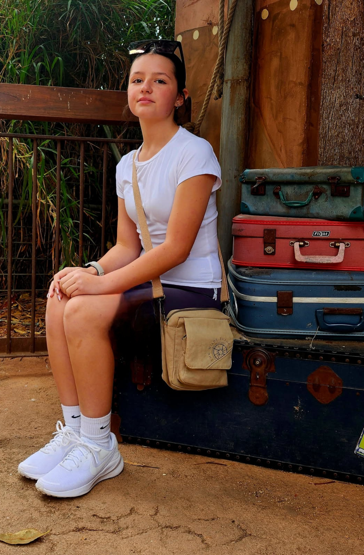 a young adventurer waits for a train sitting on a stack of battered old suitcases, with a mini canvas crossbody across her shoulder, and sunglasses ontop of her head