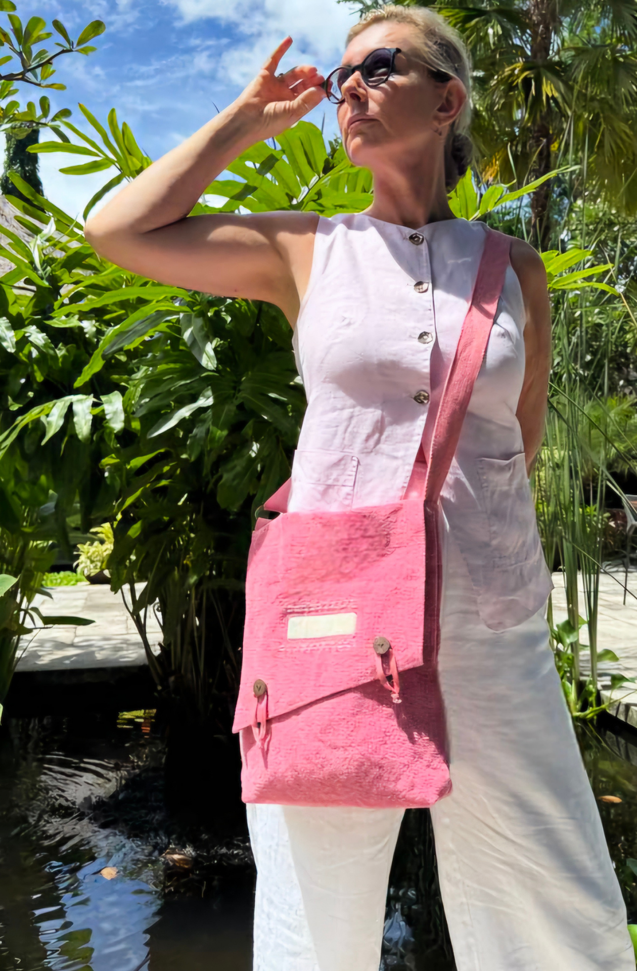 A lipstick pink jute crossbody satchel worn by a blonde traveller, with her hair in a messy bun, looking towards the sun, against a backdrop of a waterfeature with green palms'