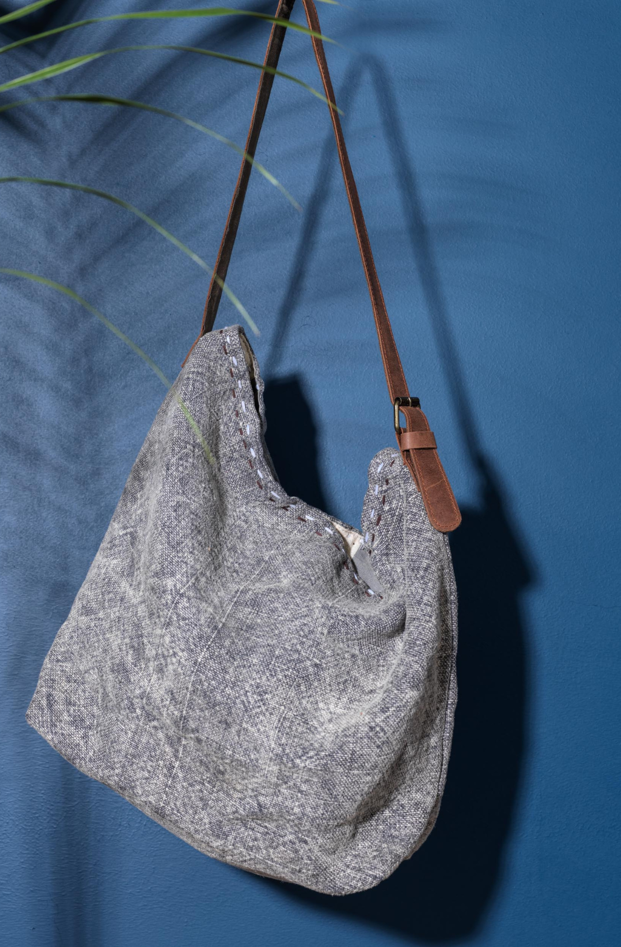 The smoke variant of the jute bag hanging from out of frame against a blue backround next to a green leafy plant.