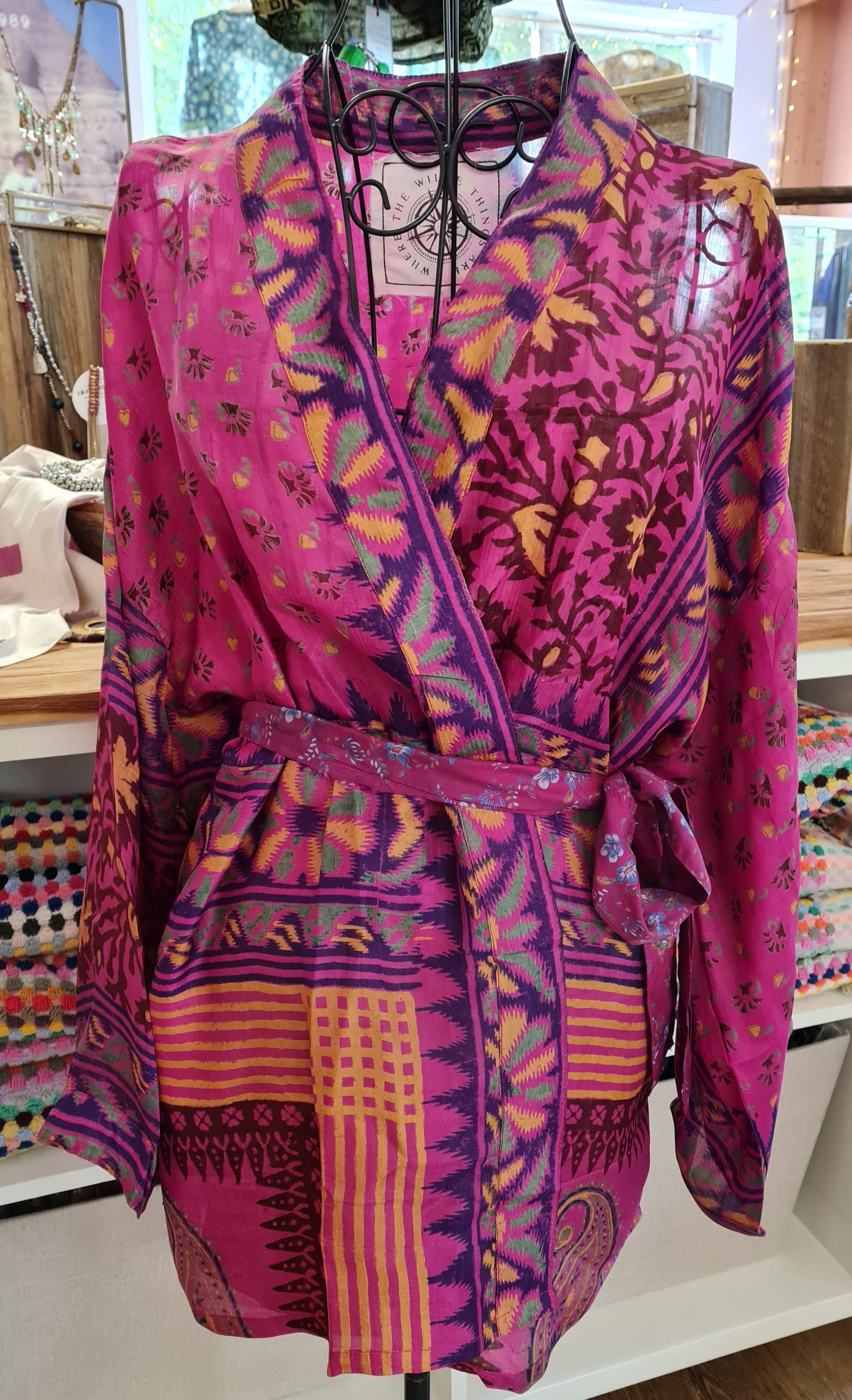 A vibrant pink kimono is displayed on a wire mannequin. The jacket has four panels of patterns, the top two are both a floral pattern, the left a simplistic and wide spread small flowers, the right shows a bold maroon and yellow winding vine floral pattern. The bottom panel is a various geometric and organic patterns in purple, yellow and a deeper pink. The fourth panel lines the sleeves and front of the jacket, a bold chunky geometric flower pattern.