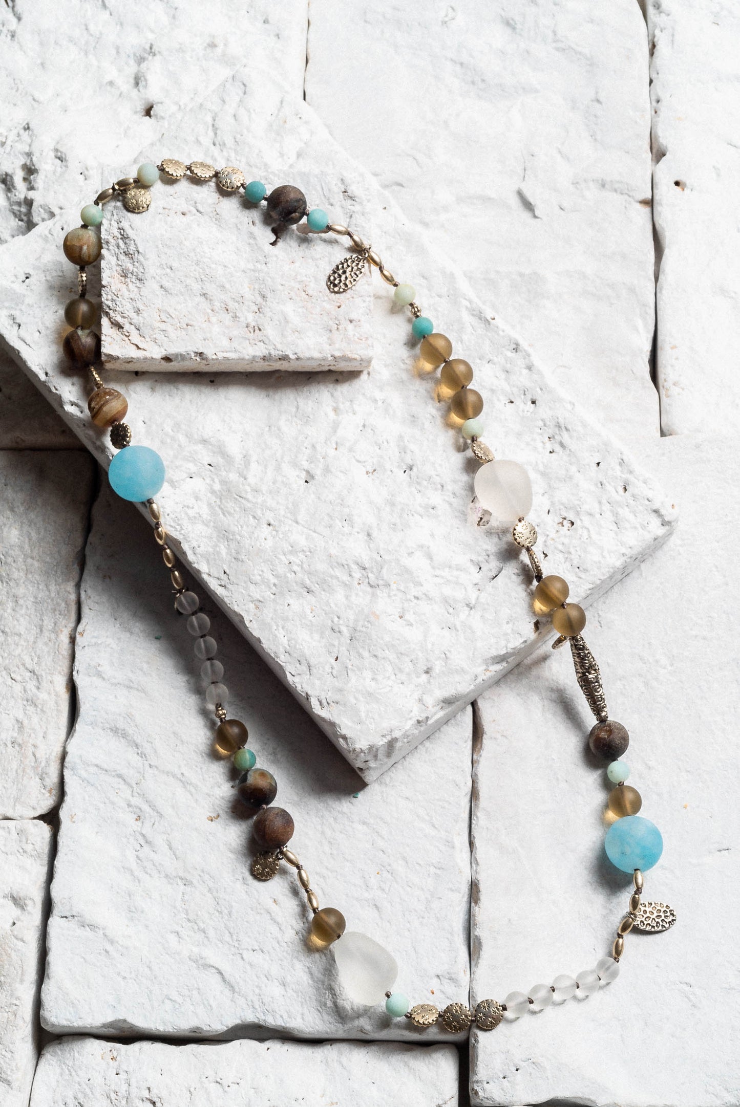 a long crystal beaded necklace. Featuring a variety of coloured and sized beads, brown, blue, grey, and white. Small silver leaf charms dotted around the necklace