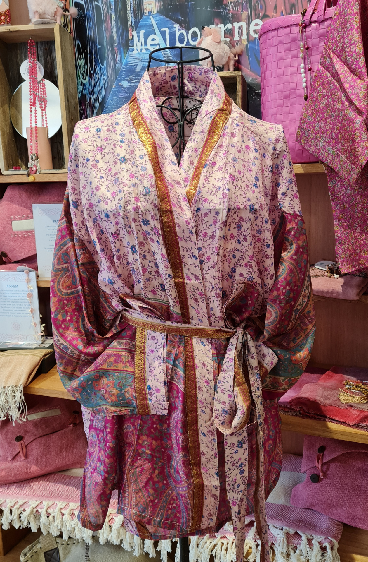 A busy patterned pale pink kimon jacket displayed on a wire mannequinn. The body of the jacket is pale pink with a tight knit blue and pink floral pattern. The sleeves look like beautiful wallpaper/traditional painting patterns, in deep warm pinks and turquoises. There is a gold trim on the edges of the jacket