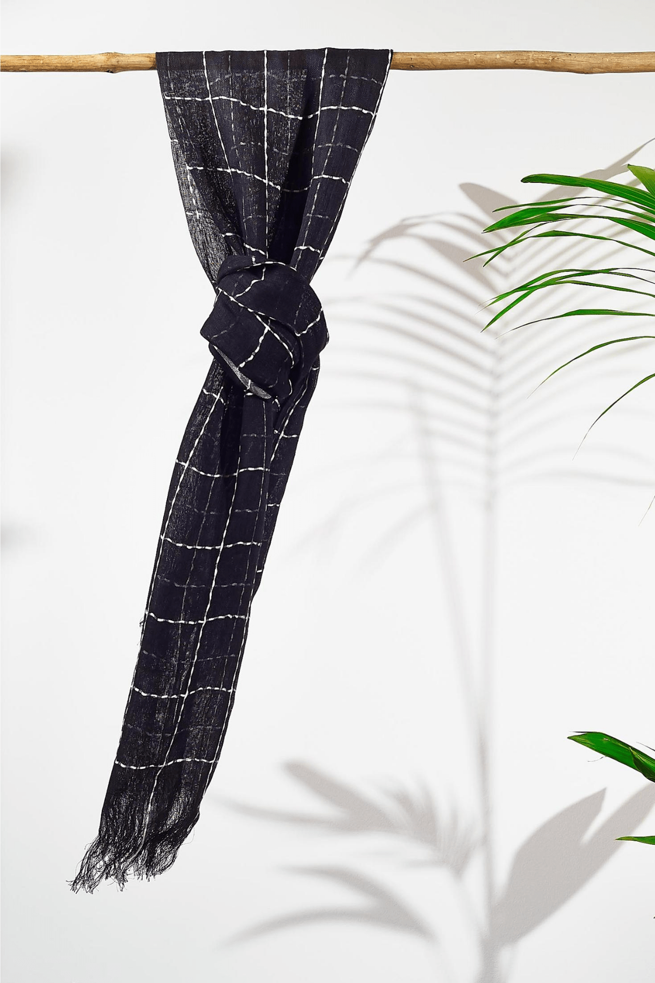 black linen scarf tied in a knot over a natural wooden rod on a white background next to a plant. Showing a different way you could tie the scarf around your neck as opposed to just draping it.