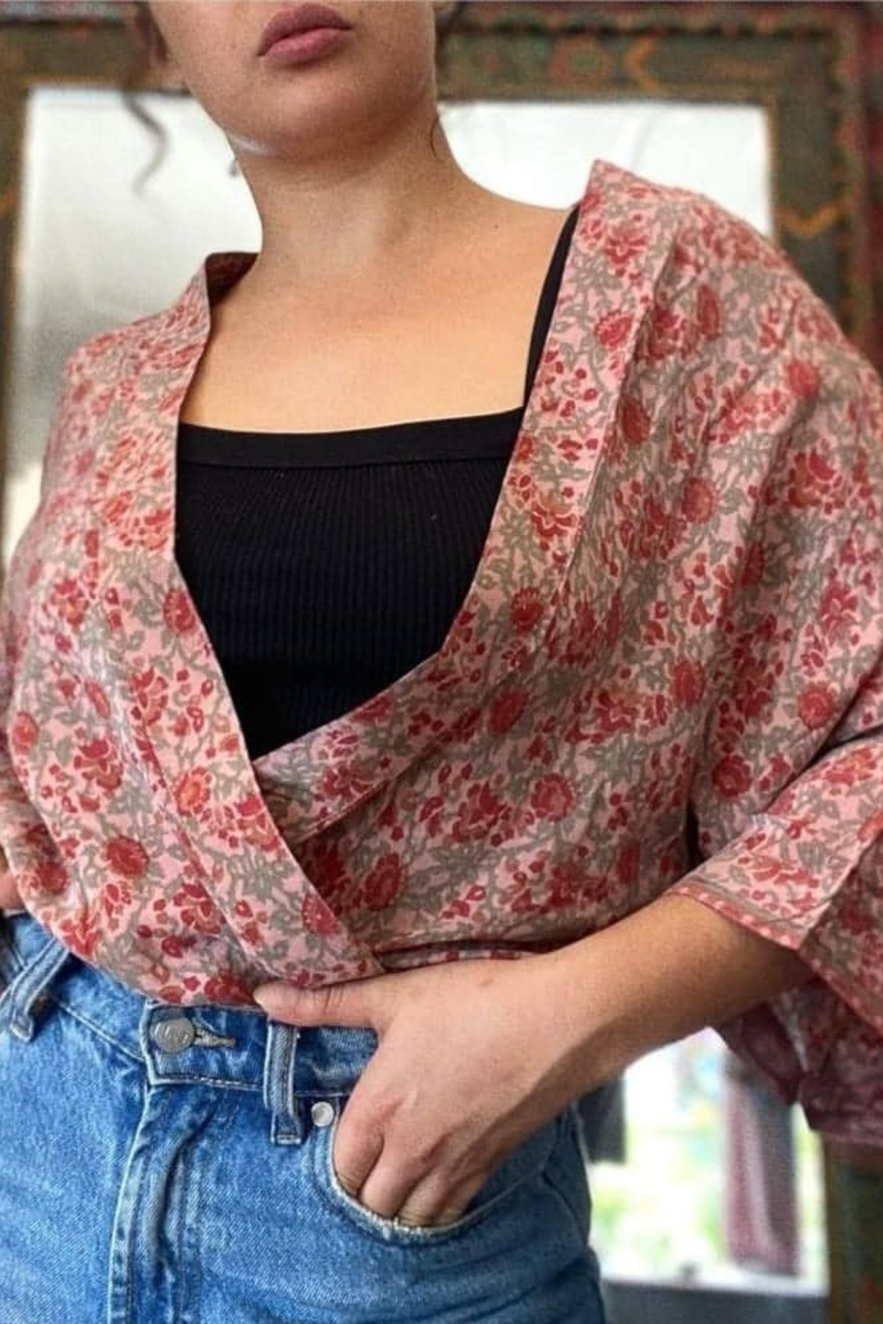 A lady stands with her hand in the pocket of her blue jeans. The shot is from the mouth downwards to the hip highlighting the muse jacket she wears on her shoulders. The jack is a dusty pink with 3/4 sleeves. A dashing red and grey-pink floral patterns covers the whole jacket.