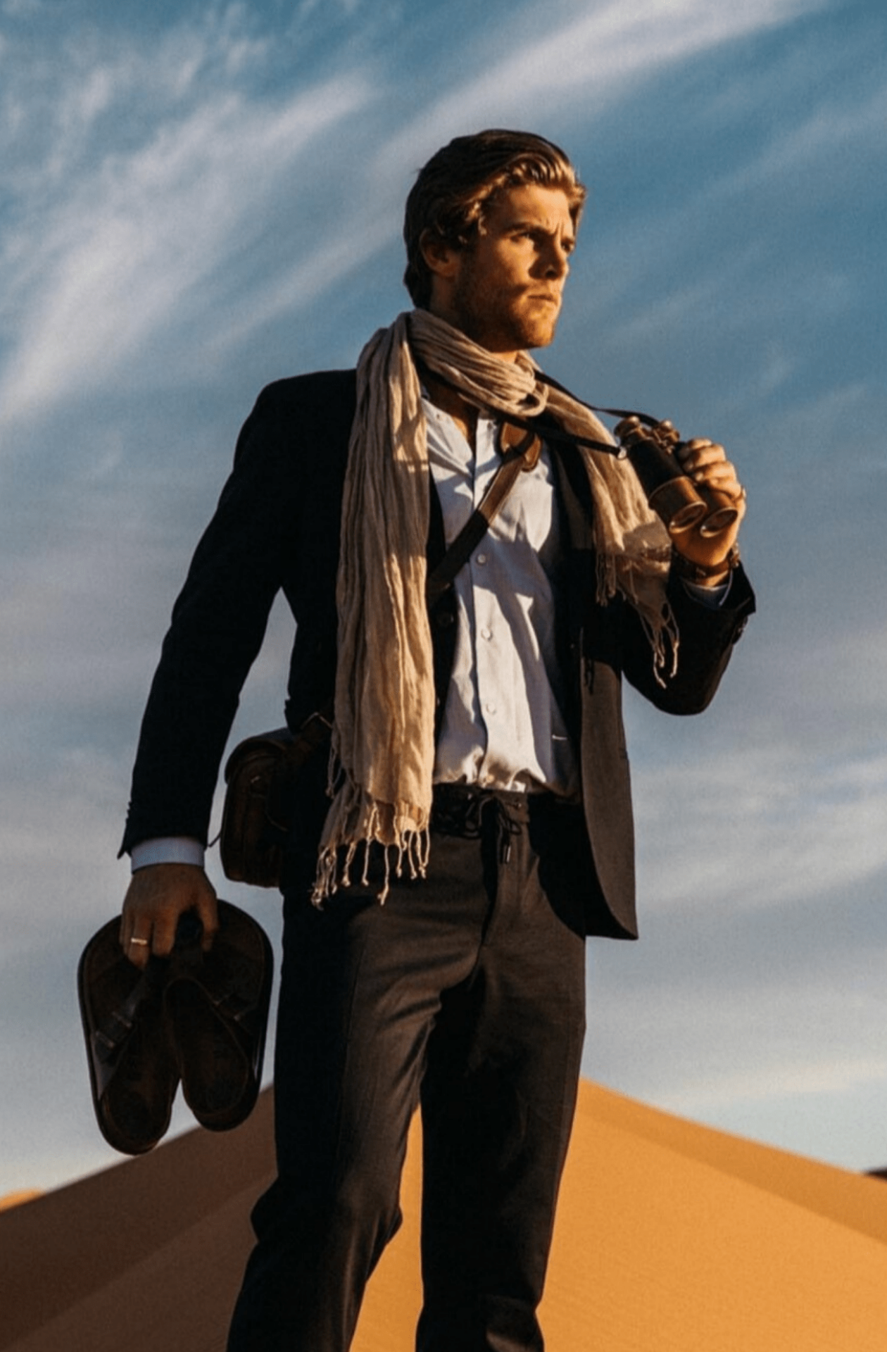 Jungle Moss scarf around a mans neck, he wears a suit and a camera bag with vintage binoculars in his hand. He is holding his shoes in the other hand and standing in a desert against a bright blue sky with wispy clouds