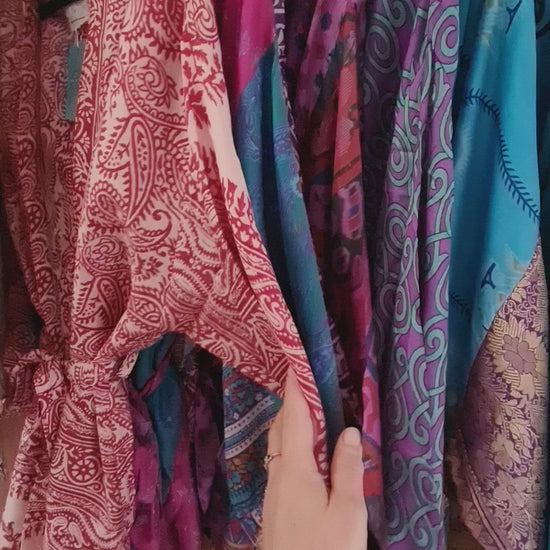 The video shows a feminine hand running along a display of silk kimono jackets, showing individual patterns available as well as the smooth silky texture of the fabric.. The jackets are aligned in colour order.