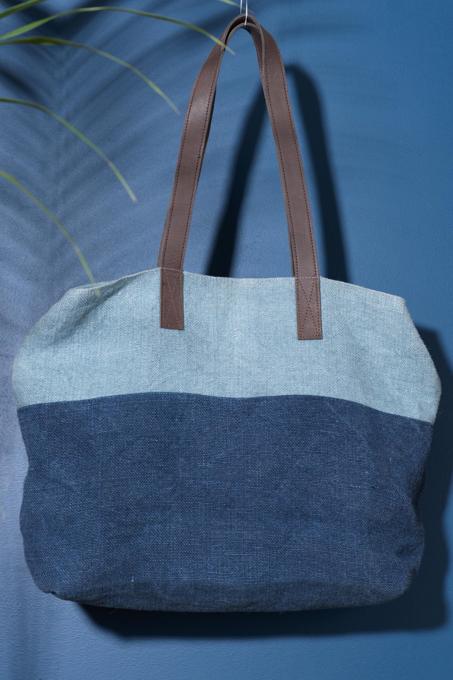 A large over the shoulder bag. With warm deep brown leather straps, and a two toned denim body. A light blue denim thick strip covers 1/4 of the bag and the rest is a classic deep blue denim. Against a blue background