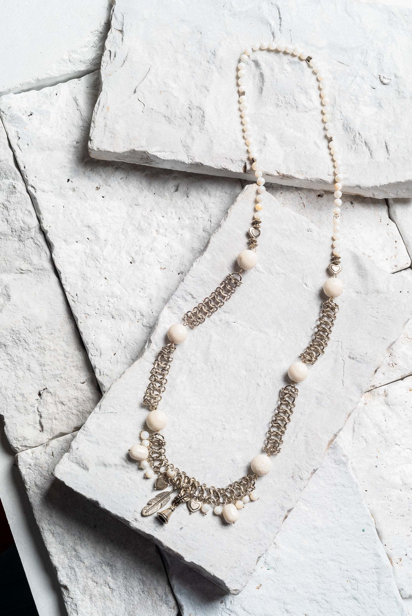A long necklace made of dainty silver chain links and large white beads. The middle features several charms, a leaf, a small bell, and two hearts.