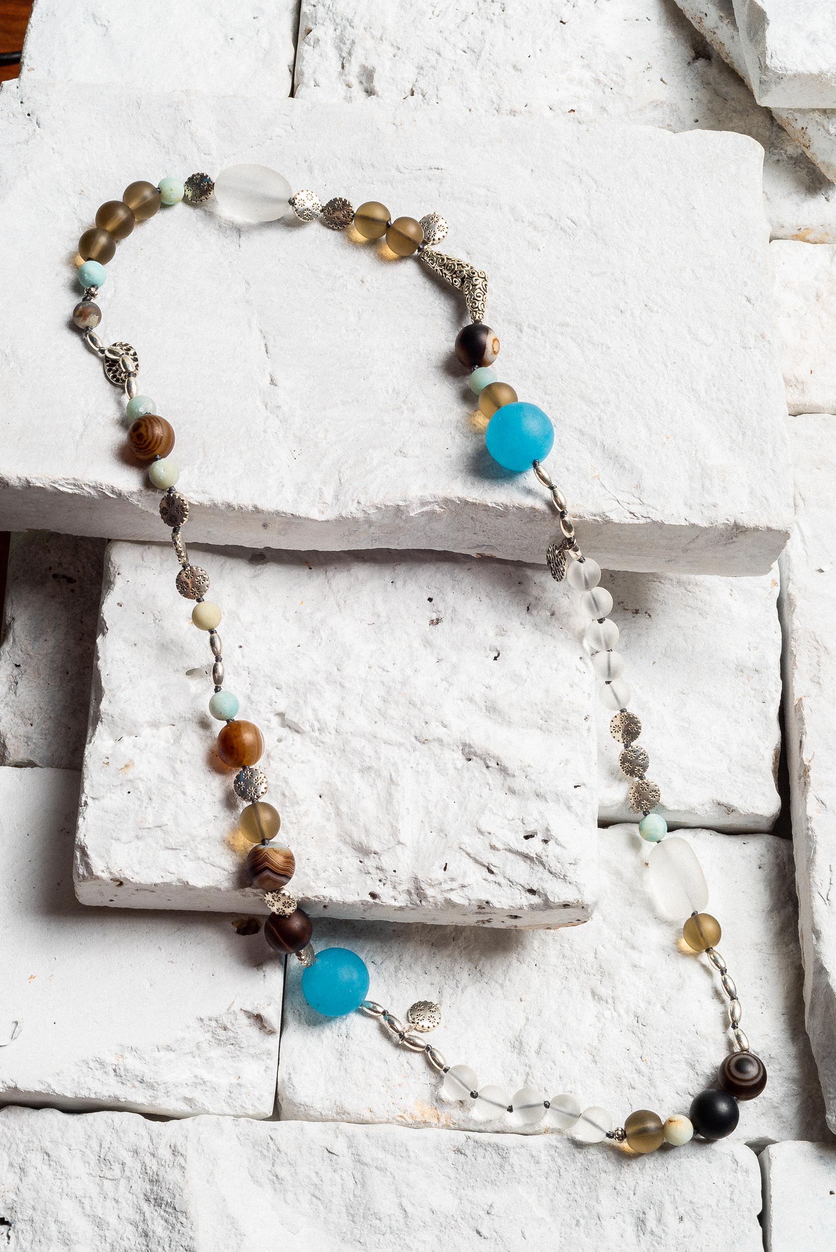 a long crystal beaded necklace. Featuring a variety of coloured and sized beads, brown, blue, grey, and white. Small silver leaf charms dotted around the necklace