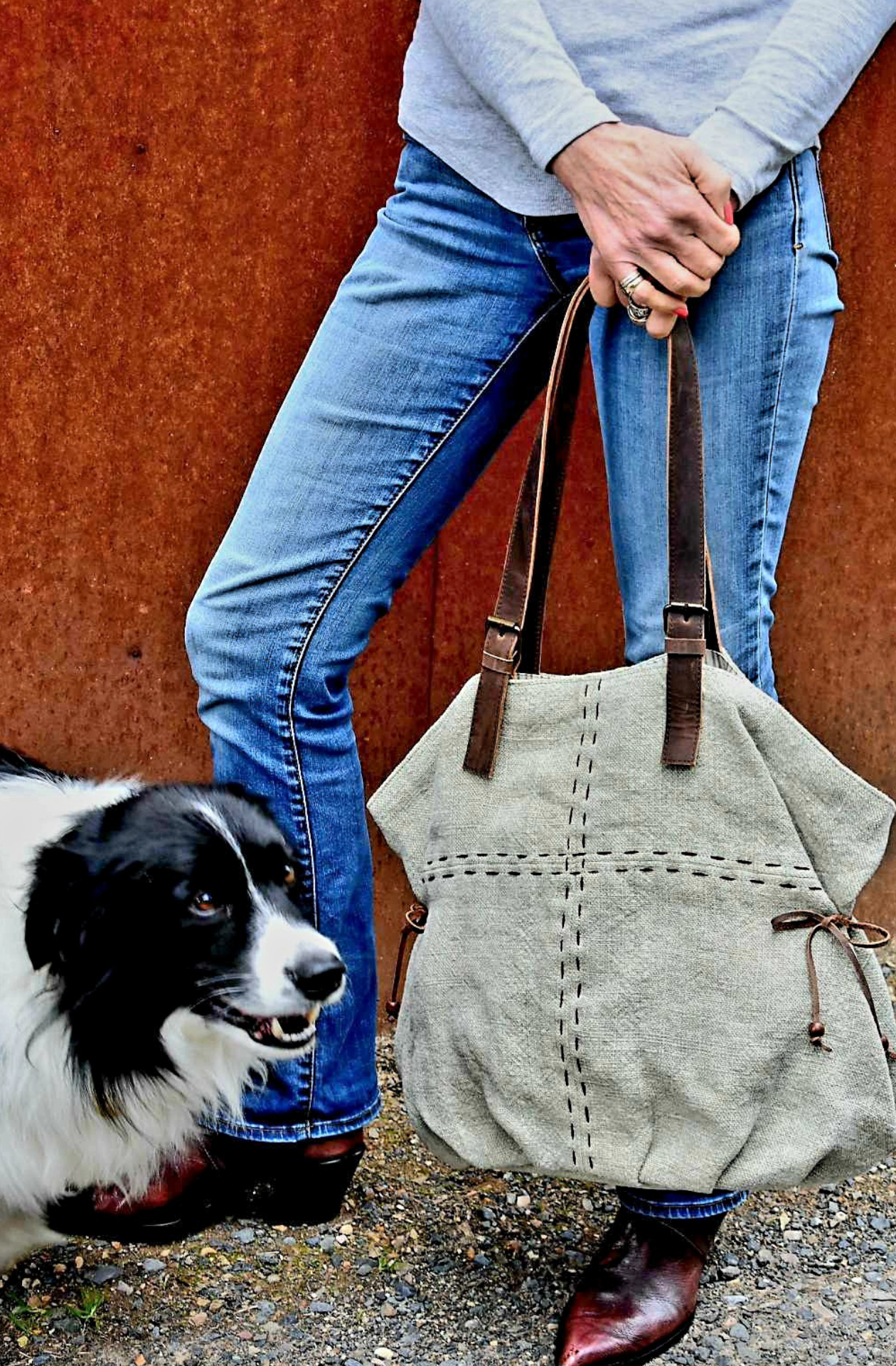 A cheeky Border Collie photobombs our model holding a large slouchy jute shoulderbag in colourway Lichen by the leather straps. She is holding the bag low showing her blue jeans and vintage cognac coloured cuban heeled boots