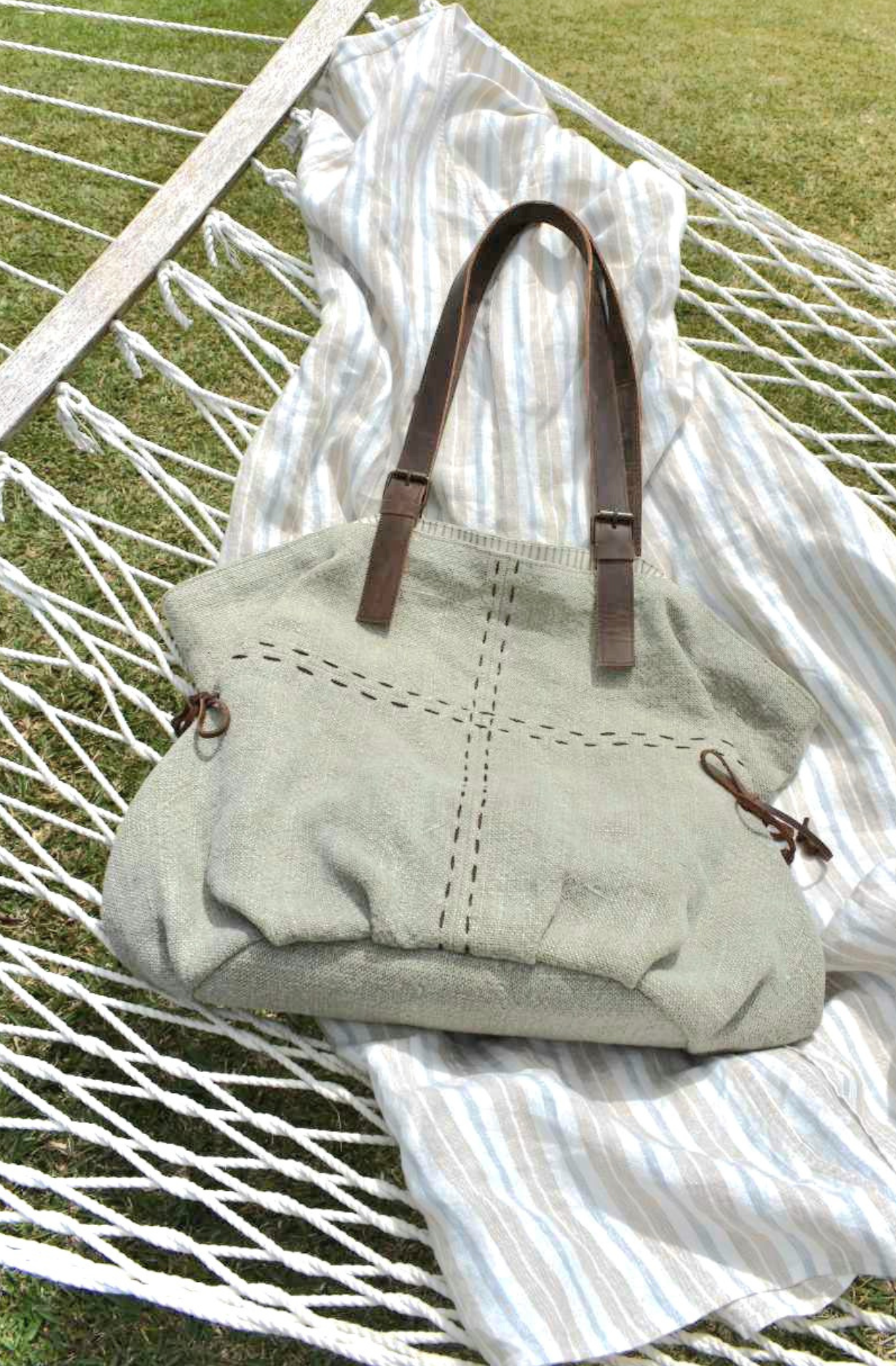 a soft green grey jute shoulder bag with both vertical and horizontal stitching through the middle lies on a linen dress on a hammock in Vanuatu  It has side ties and 2 shoulder straps in leather. The straps are joined at the front by 2 brass buckles. The striped lining is just visible at the top where the bag is slightly open. 