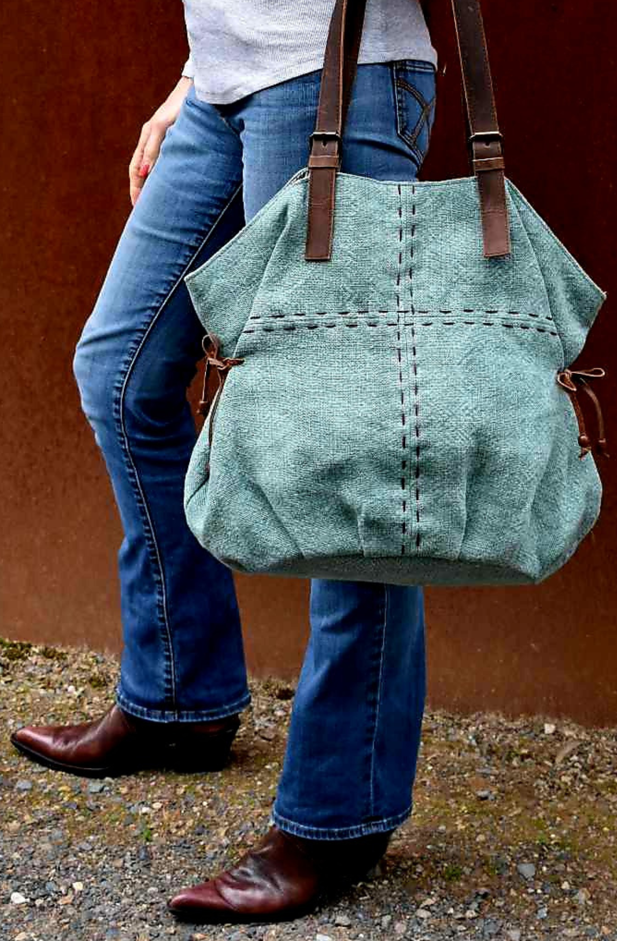 A model holds a Persian Blue jute slouchy shoulder bag across her forearront of her jeans so that it swings infront of her blue jeans, against a rusty wall. 