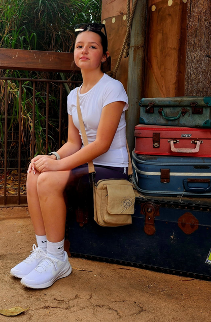 a young adventurer waits for a train sitting on a stack of battered old suitcases, with a mini canvas crossbody across her shoulder, and sunglasses ontop of her head
