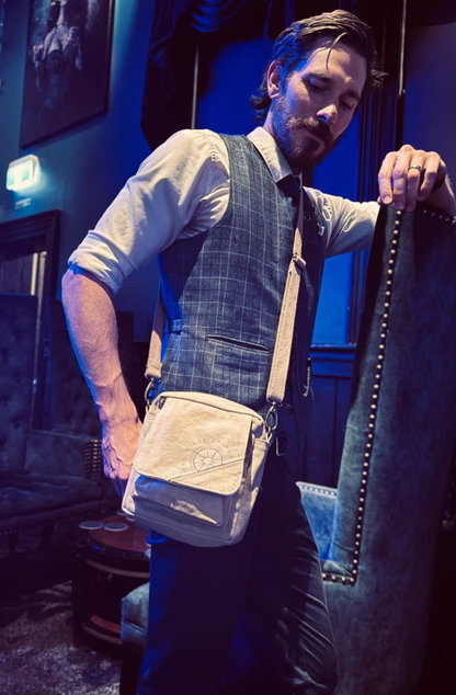a trendy well dressed guy wears an All You Need canvas crossbody against his checked blue waistcoat. He leans against cobalt blue velvet highbacked chairs Lighting is indigo blue in a Melbourne Cocktail Bar