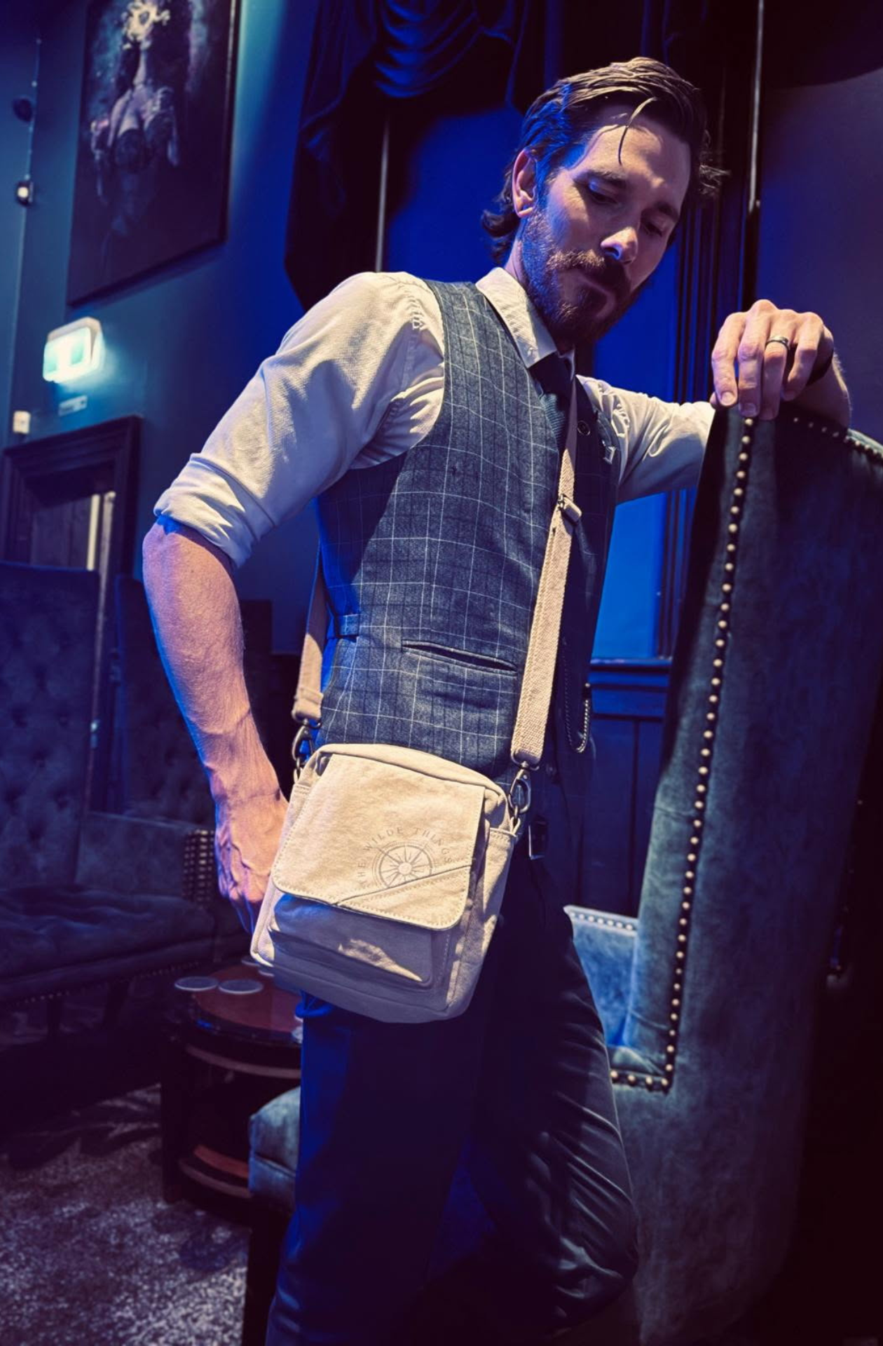A fashionable young man poses casually in cocktail bar, with a Where The Wilde Things Are sustainable canvas crossbody bag slung over his shouder
