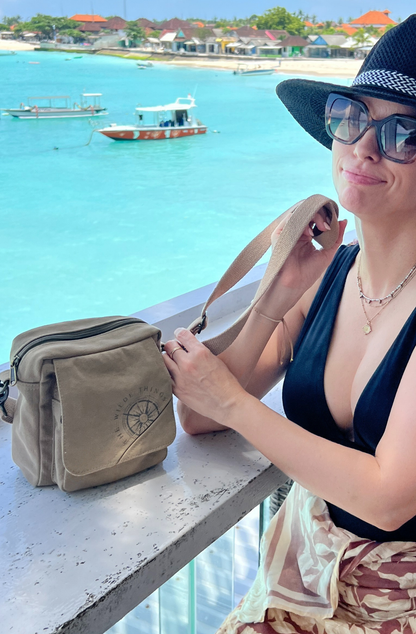 A beachside tourist wearing black hat and bathers, is picking up her All You Need mini canvas crossbody from an outdoor bar. the background is blue ocean with small boats seen just offshore. 