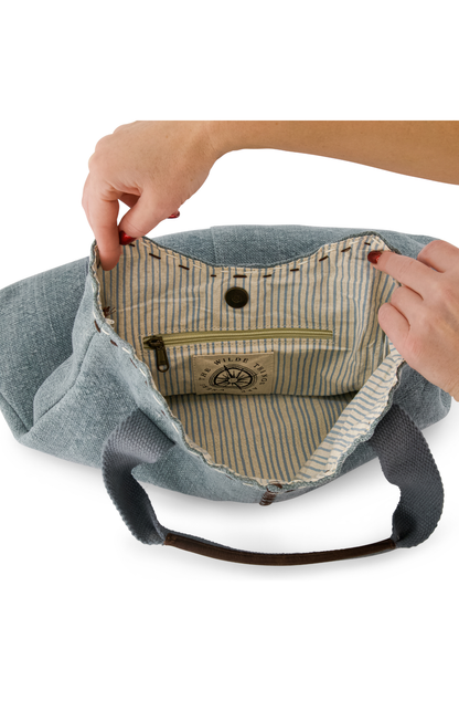 Light blue, chambray coloured jute bag showing pin striped cotton lining and interal zip pocket, hold open by a model's hands.