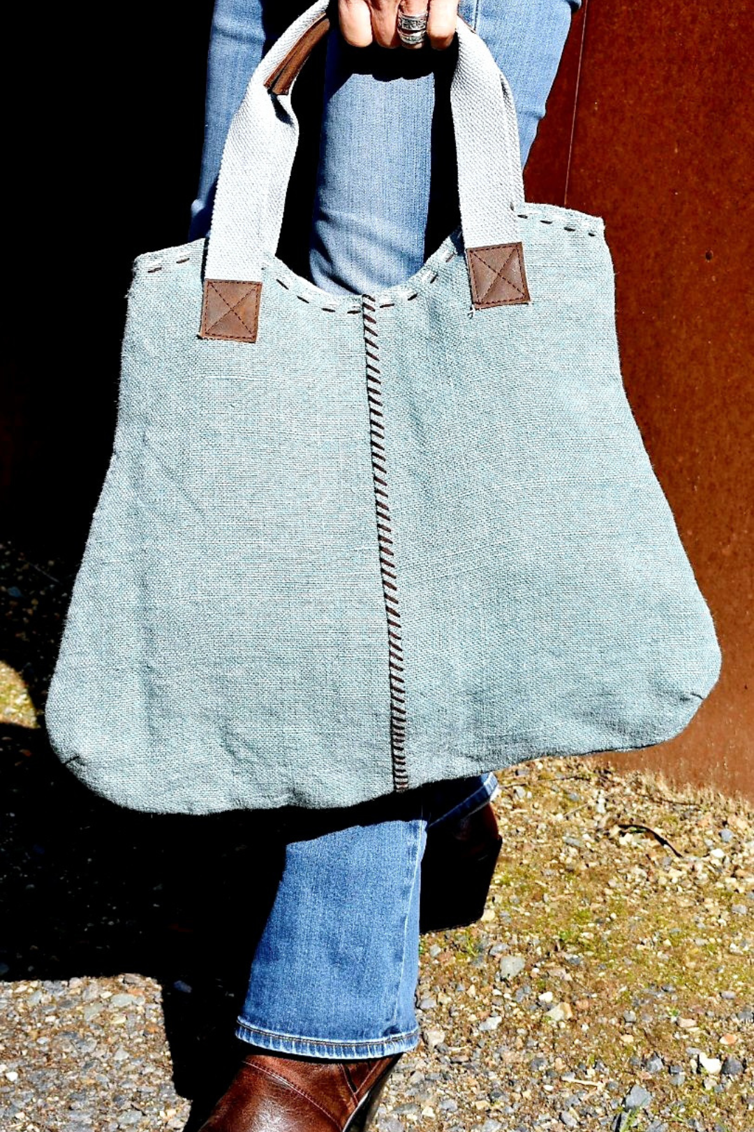 A model's hand is just visible at the top of the image holding the short straps of a faded blue jute shoulder bag, infront of her blue jeans.