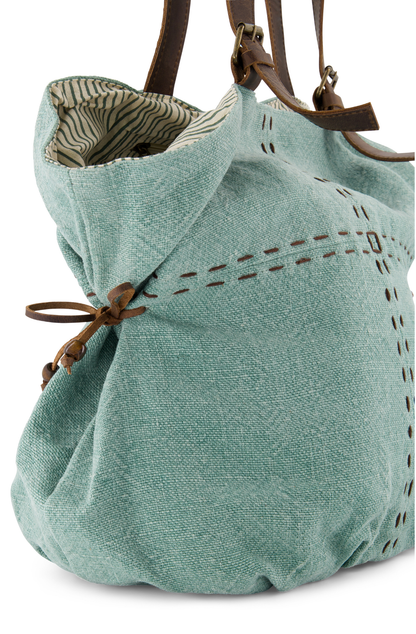 a side view of a shoulder bag with a large carrying capacity and decorative hand stitching, the shoukder bag has leather side ties to give it an appealing shape and two leather shoukder straps with decorative brass buckles. Its colour is a French Blue, made from ethically sourced jute. The pin striped lining can be seen at the top edge.