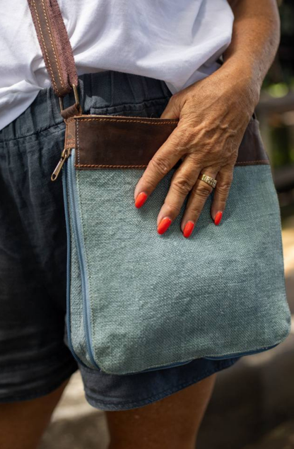a holiday maker shows a close up of a chambray coloured mini cross body bag showing the side zip open to expand the bag.  er hand, with coral coloured nail polsh, hols the top edge of the bag.