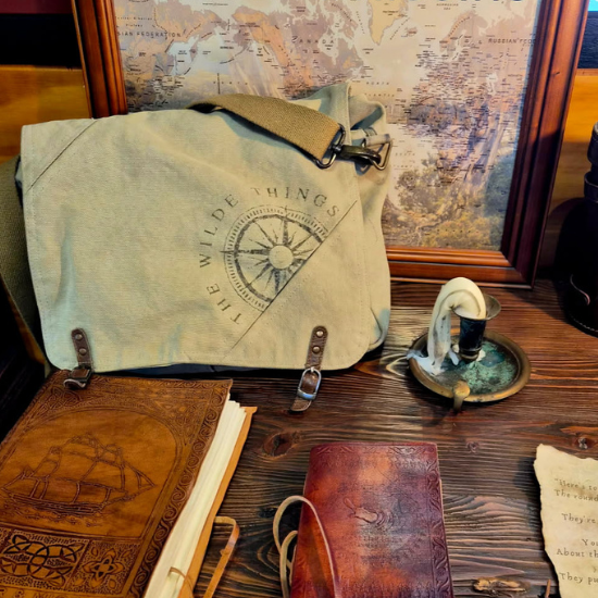 Canvas bag with 'The Wilde Things' compass logo on an antique wooden desk  with two leather covered journals  an olde world map in the background