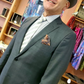 A portrait of a man with a deep grey suit on, a brown and orange pocket square in his breast pocket