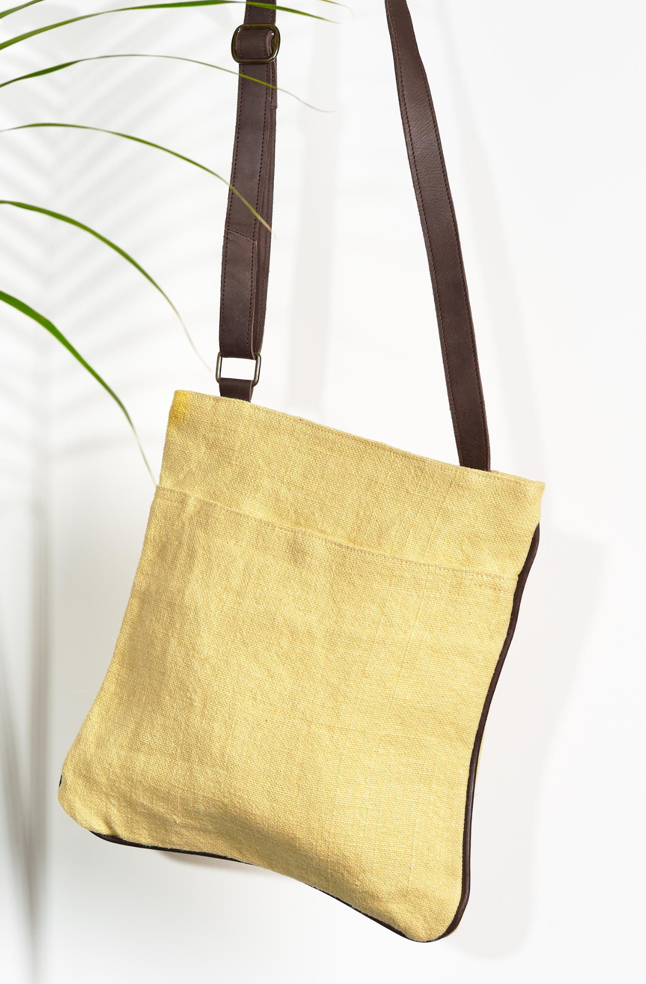 The "Daffodil" variant of the Hold Me Close Cross Body Bag. It is a baby yellow with an open front pocket, with matching brown leather piping around the outside edge. An adjustable dark brown strap is connected on either side of the bag.