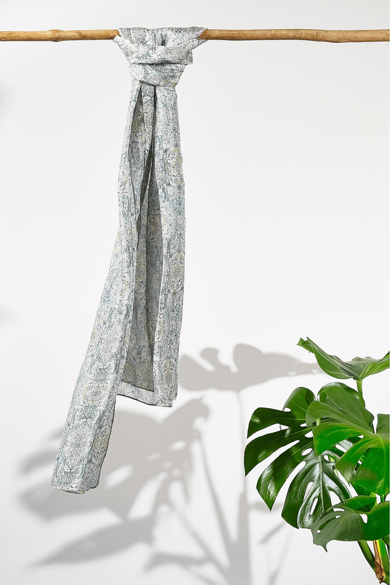 The pistachio moroccan silk scarf hanging from a rod. Showing how smooth the silk is as it hangs. The scarf is a pale grey with a deep grey-green busy pattern on it