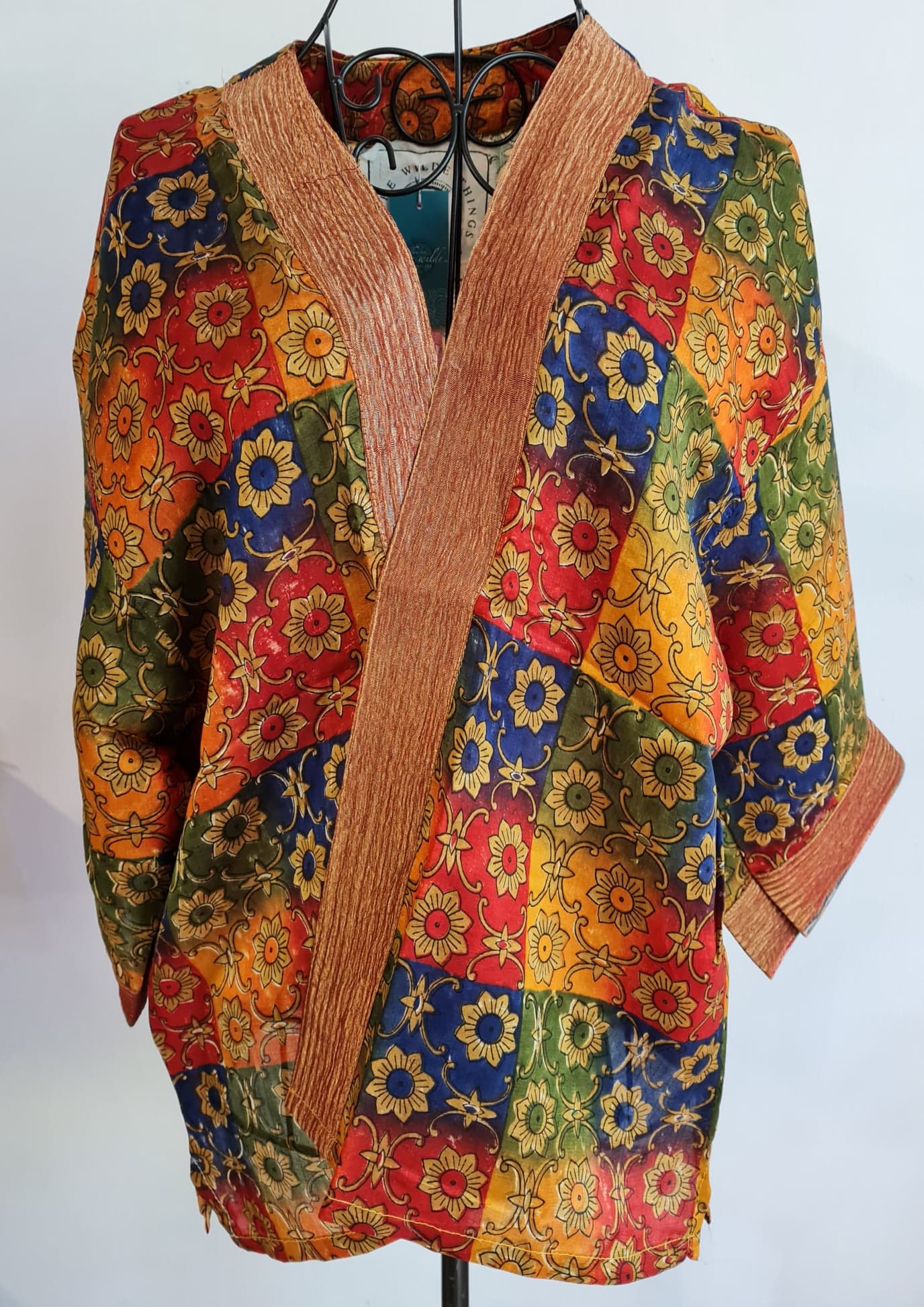 Regalia muse jacket crossed over a wire mannequin, squares in blue, green, red and yellow make up the jacket. On top of the squares are a light golden floral pattern in a geometric fashion. the jacket has a textured red silk trim. Regalia