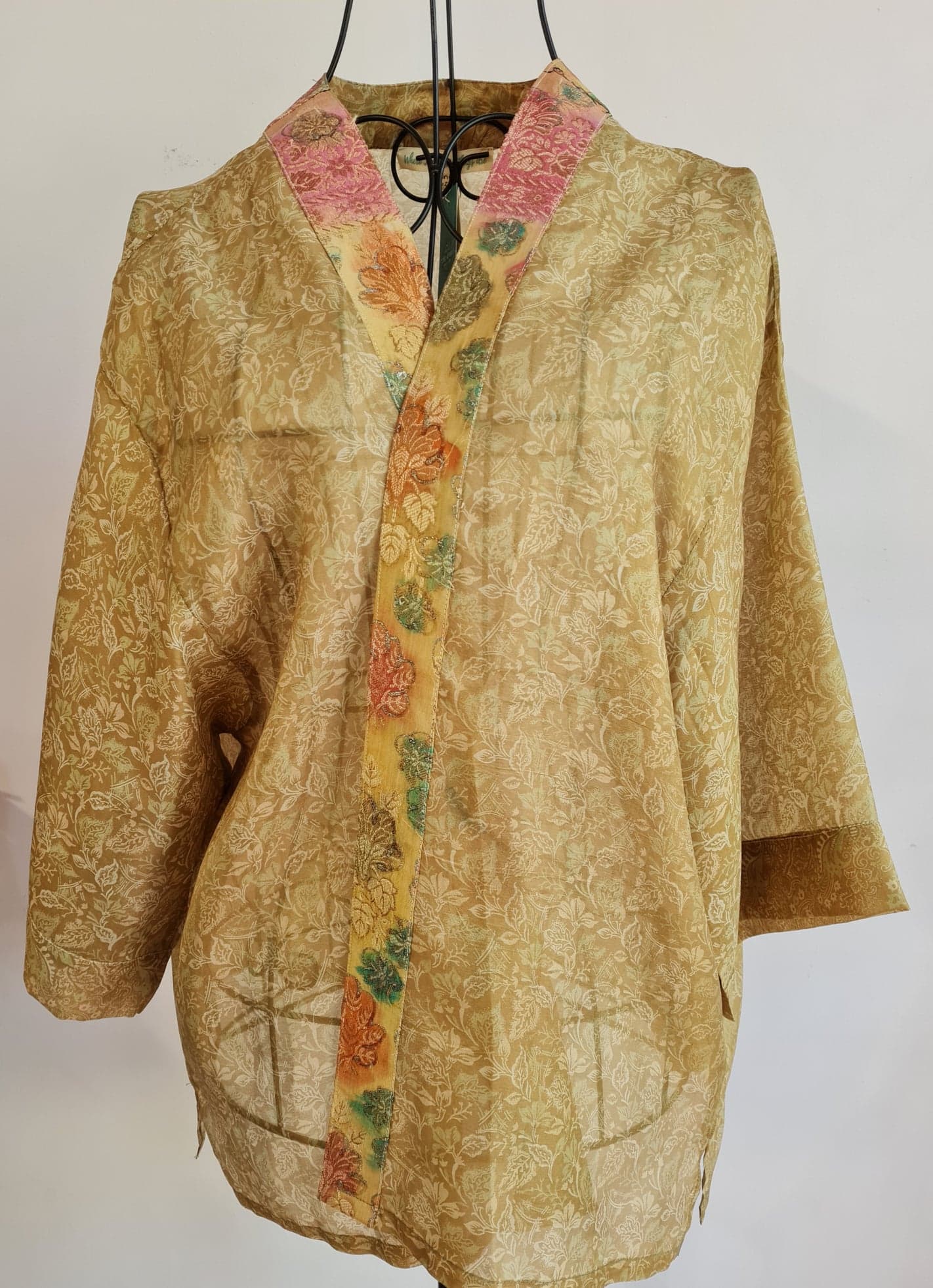 a cool yellow muse jacket is crossed over a wire mannequin. It features a pale yellow floral pattern covering the whole thing. a deep yellow trim lines the jackets front, with warm oranges, yellows and green leaves watercolour styled pattern, the trim itself gradients upward into a light red colour toward the neck of the jacket. 