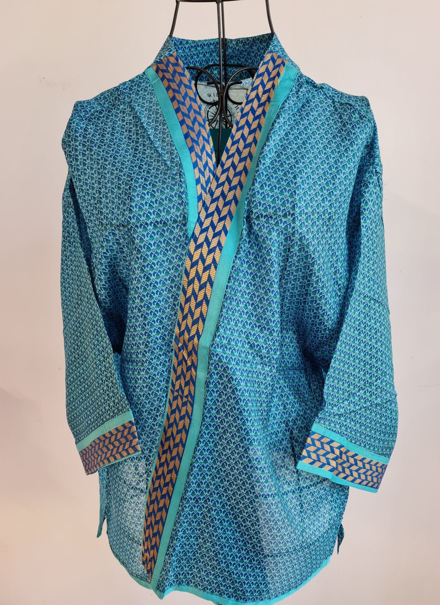 a busy floral turquoise muse jacket is crossed over a wire mannequin. It features a variety of shades of blue and a golden blue trim around the front