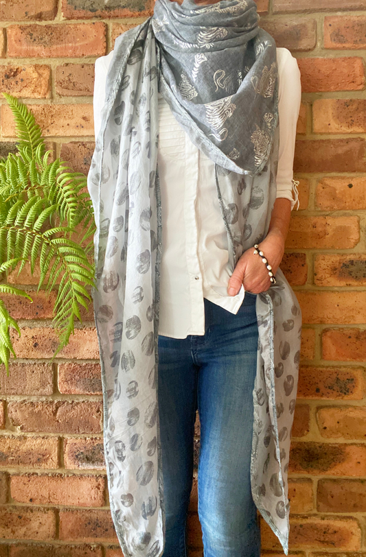 A woman with the Forests of the Night scarf wrapped around her shoulders like a shawl. The scarf is a silvery grey with reflective silver tiger patterns on the cotton section and medium grey dots like full moons screen printed on the silk ends. 