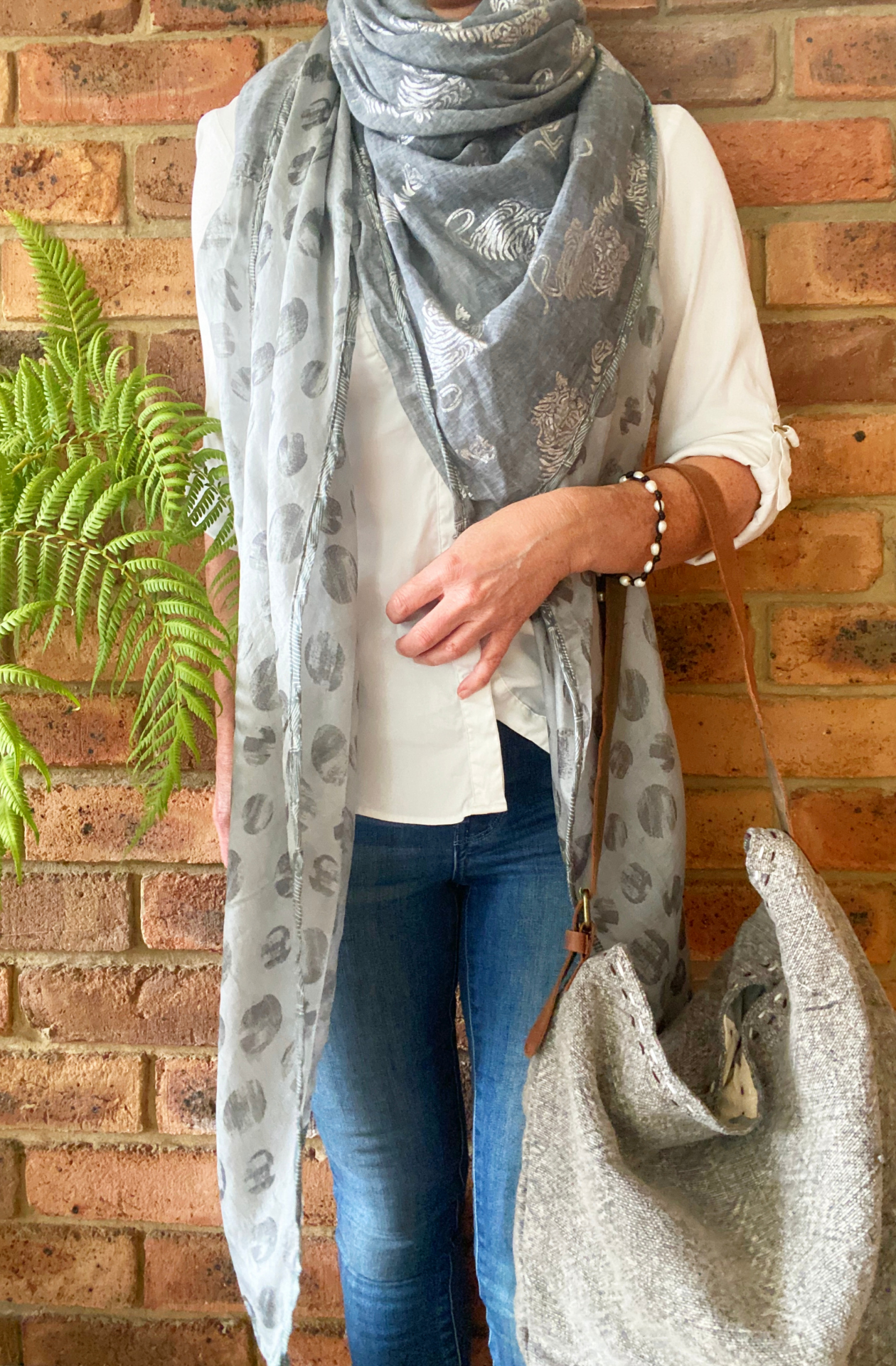 A woman with the Forests of the Night scarf wrapped around her shoulders like a shawl. The scarf is a silvery grey with reflective silver tiger patterns on the cotton section and medium grey dots like full moons screen printed on the silk ends. Over her arm is a Smoke grey, jute shoulder bag called All The World.