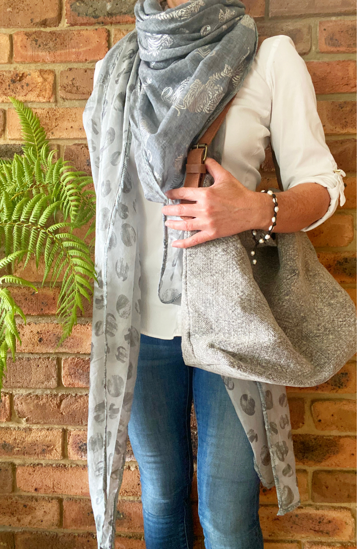 A woman with the Forests of the Night scarf wrapped around her shoulders like a shawl. The scarf is a silvery grey with reflective silver tiger patterns on the cotton section and medium grey dots like full moons screen printed on the silk ends. Over her shoulder, she carries a Smoke grey, jute bag called All The World.