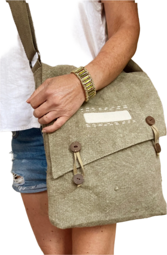 A tanned woman holds a khaki coloured stonewashed jute crossbody bag against her left hip, showing her denim shorts, white linen sleeveless top and olive green enamelled cuff bracelet. It has tab closures of 4 coconut wood buttons and cotton ties, a front decorative panel in natural coloured cotton with hand stitched highlights and is vegan friendly.