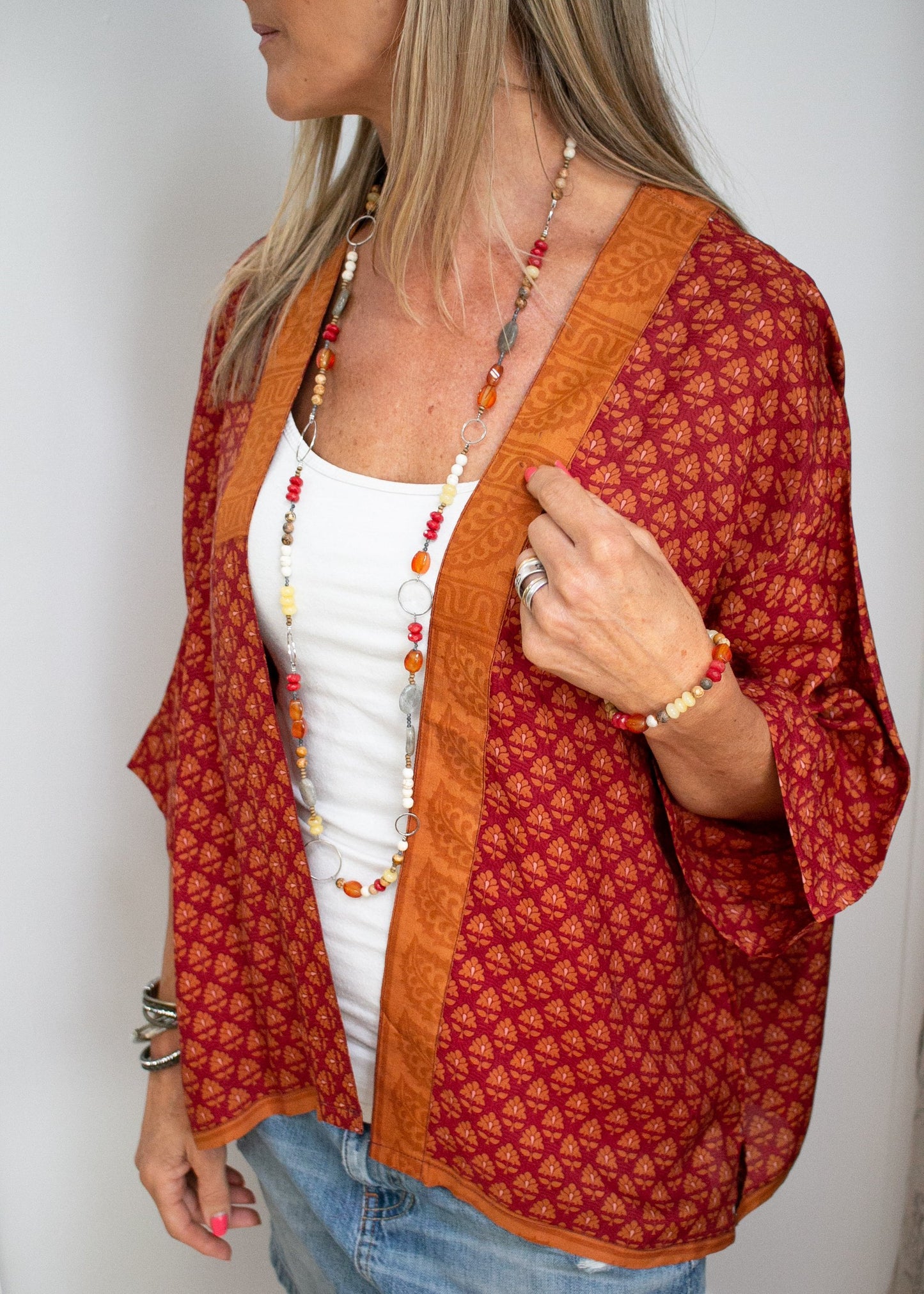 a woman wearing a deep red muse jacket with a repeating geometric floral pattern in a paler red. the trip around the front is a bold orange. She pairs the jacket with match orange and red jewellery 