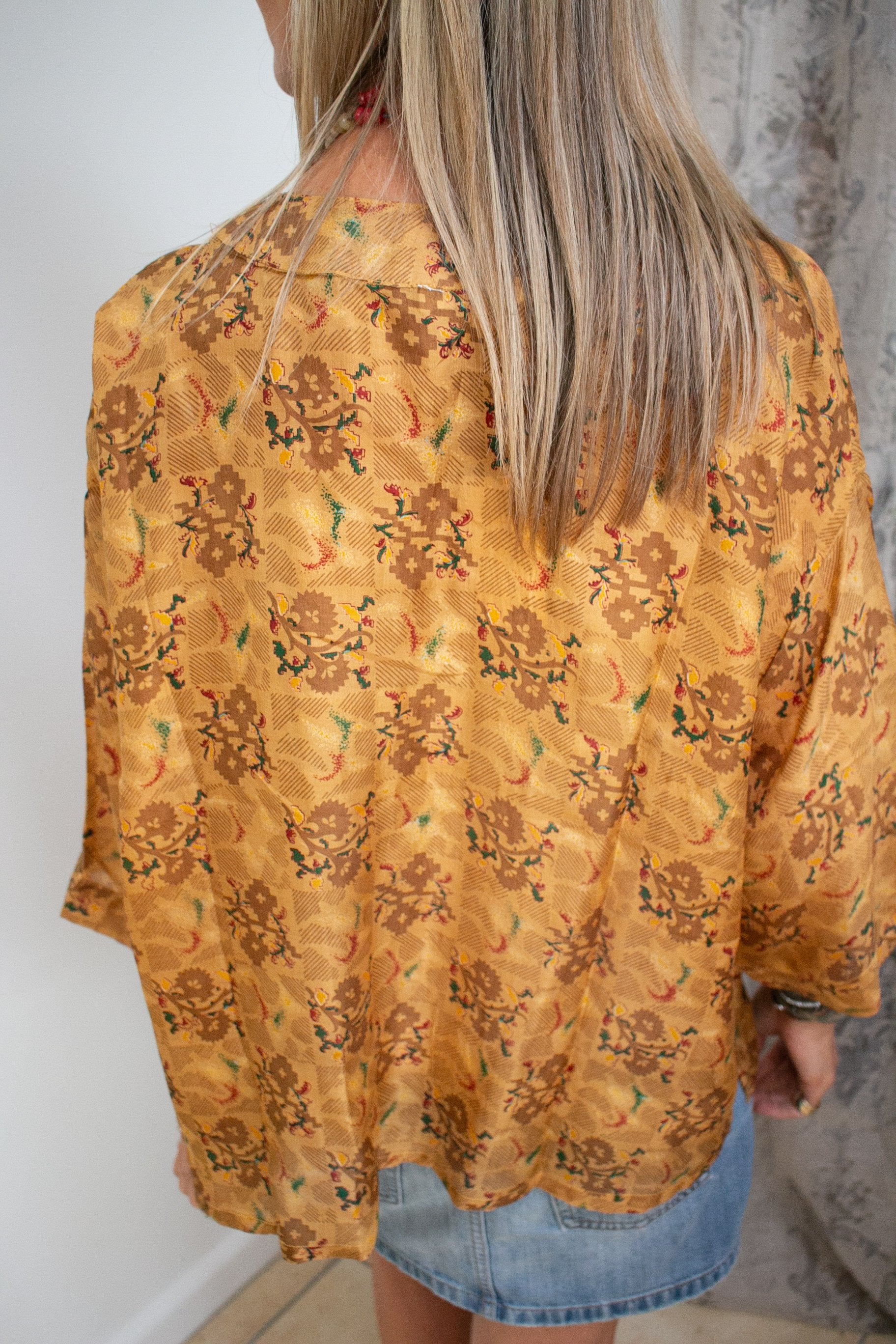 a muse jacket over a womans shoulders who is facing away from the camera to show the relaxed boxy fit of the sari silk, as well as the 3/4 sleeves. The jacket is yellow with green, red and deep yellow pattern across it.
