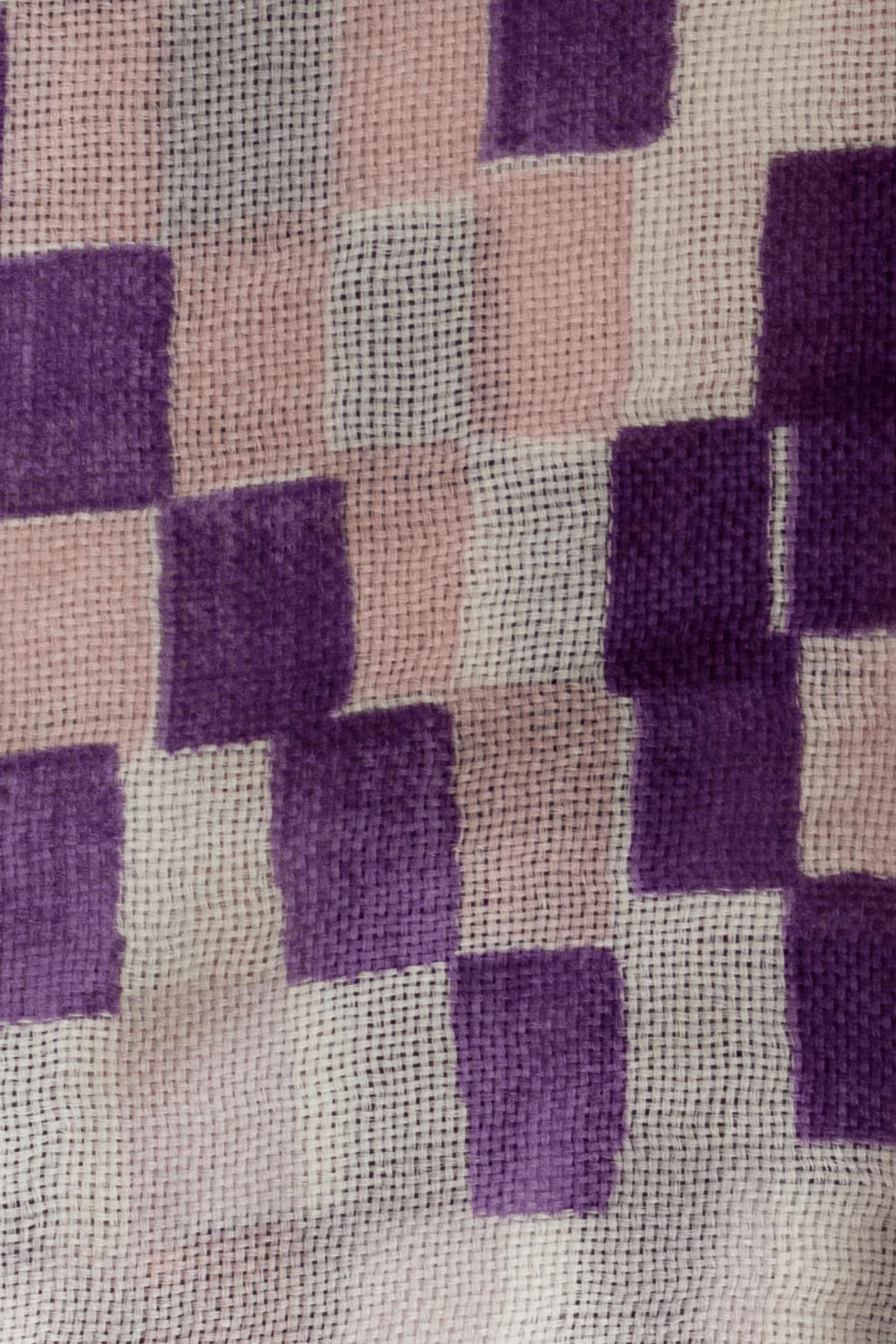 a close up of the Amethyst variation of the chequered love wool scarf. The scarf features an eclectic pattern of squares that appear painted in a variety of shades. Deep royal purple, a medium mauve shade, a pastel pink, and the white linen of the scarf. 