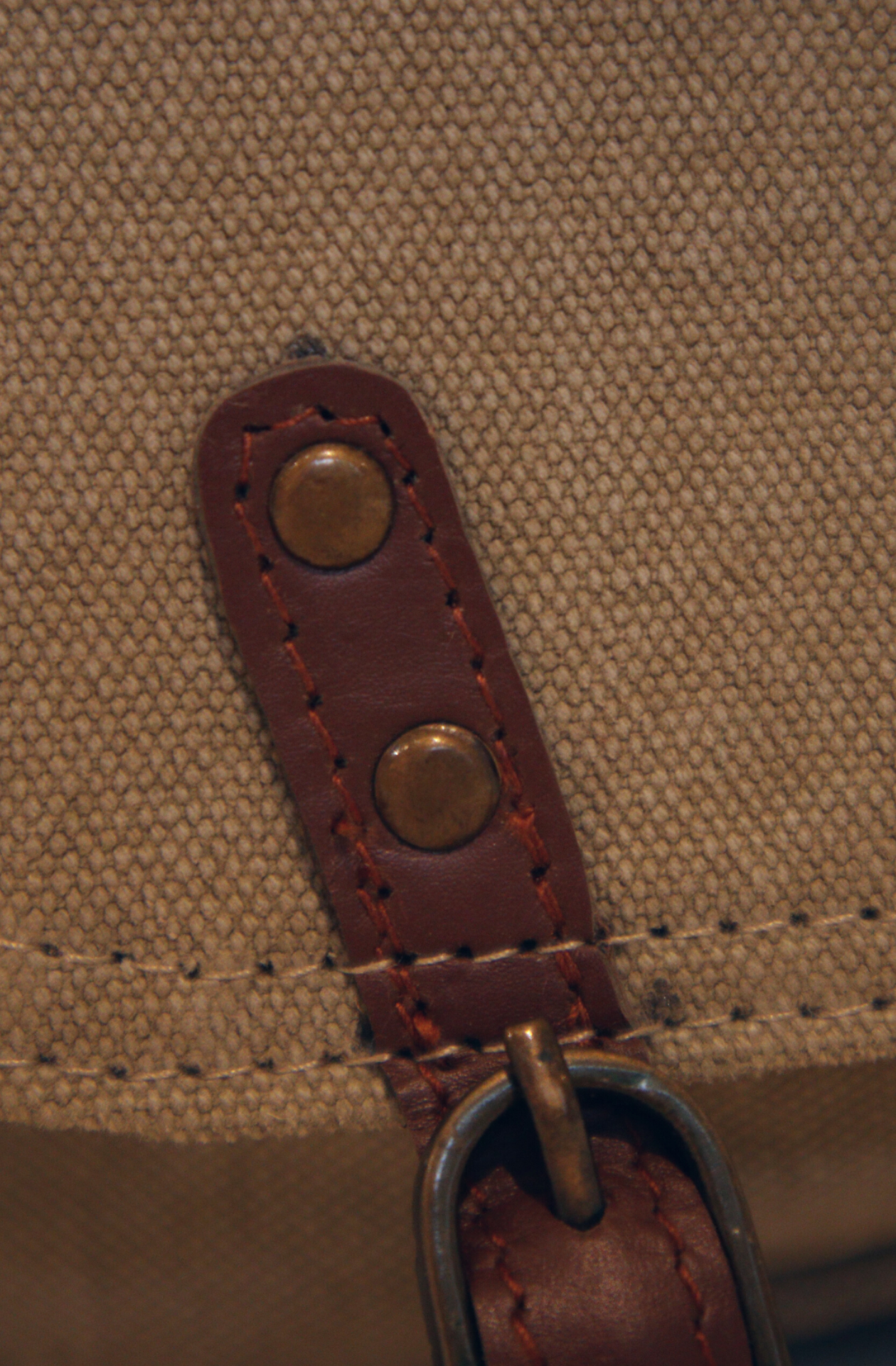 a closeup of the leather tab and brass buckle on the khaki canvas front flap of the crossbody bag named The Extra Mile.  two brass studs can be seen, which fix the tab in place