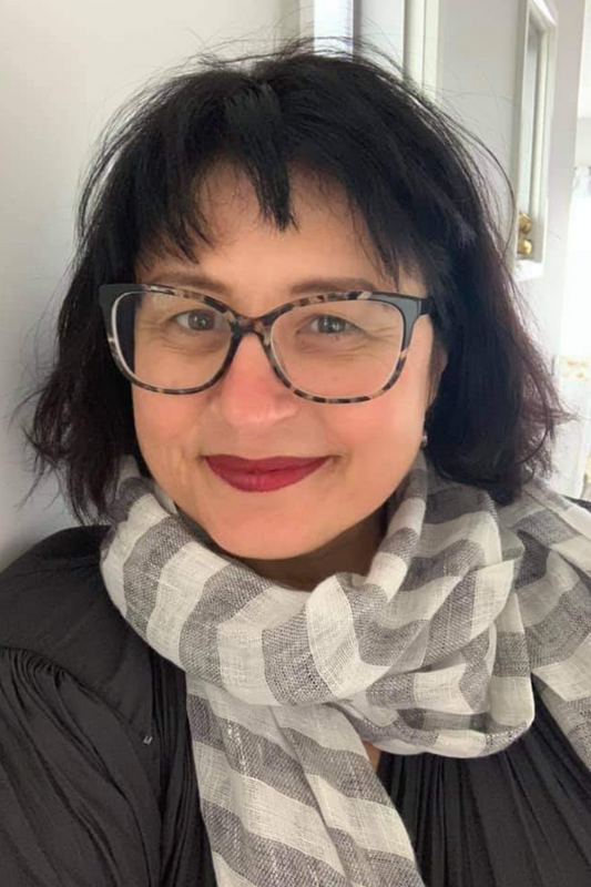 A portrait of a woman wearing large frame glasses and red lipstick. She has a grey and white vertical striped hamptons linen scarf around her neck.