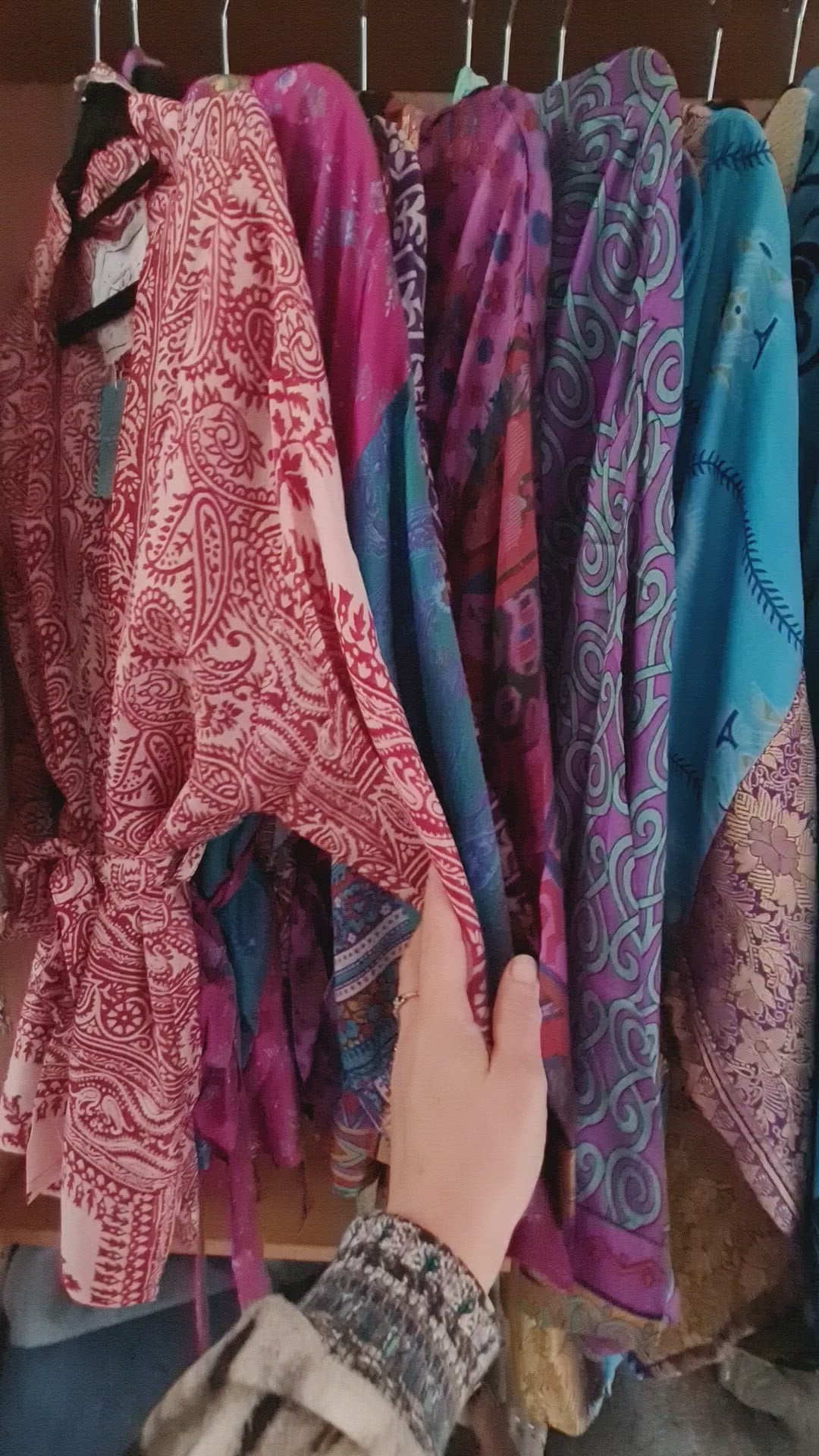 The video shows a feminine hand running along a display of silk kimono jackets, showing individual patterns available as well as the smooth silky texture of the fabric.. The jackets are aligned in colour order.