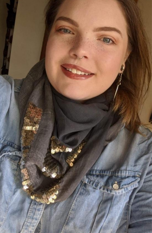 A woman wears the after 5 scarf around her neck with a denim jacket and a sword earring. She styles the scarf like a bandana and has it tucked in the front of her shirt. The scarf is a deep grey with golden sequen details