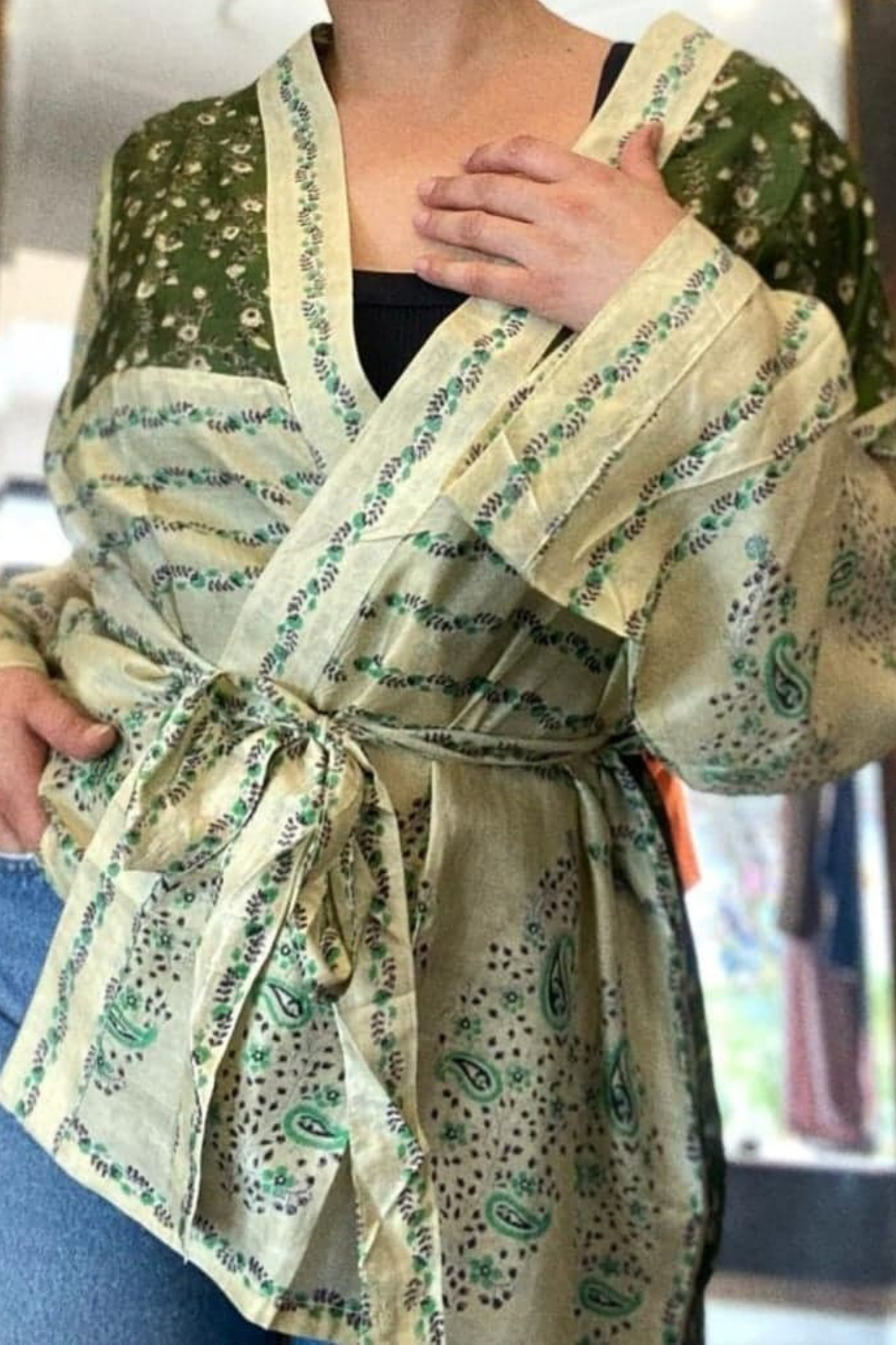 The subject is a womans torso wearing a green and off white floral/paisley kimono jacket made from vintage sari silk. the jacket is crossed across her abdomen and tied around the waist with a matching off white, green paisley tie. the woman is wearing a simple black tshirt and blue jeans underneath