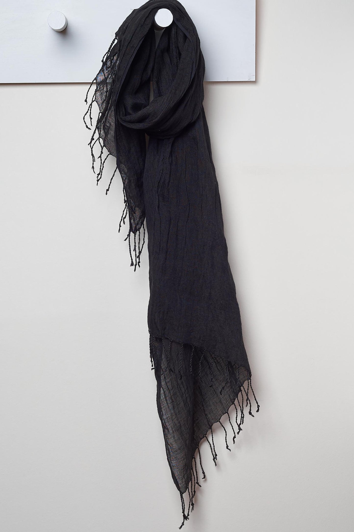 black 3 visits to cairo linen scarf.  looped around a hook detailing the linen texture and thin tassels tied at the short edges of the scarf.